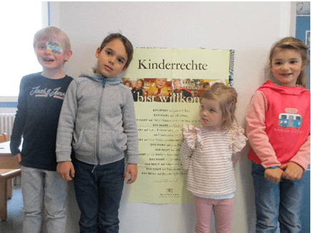 Vier Kinder stehen vor einem Plakat, auf dem zu lesen ist "Kinderrechte du bist willkommen"