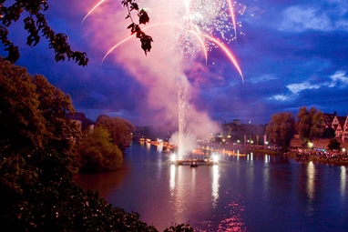 Ein pinker Feuerwerkskörper entzündet sich in gleißendem Licht über dem Wasser.