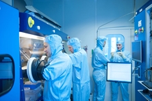 Vier Männer in blauen Schutzanzügen stehen im Labor, zwei von ihnen greifen mit Schutzhandschuhen in einen durch Glas abgedichteten Bereich.