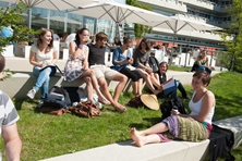 Studierende sitzen auf dem Rasen und auf Treppenstufen auf dem Campus, trinken Kaffee und unterhalten sich.